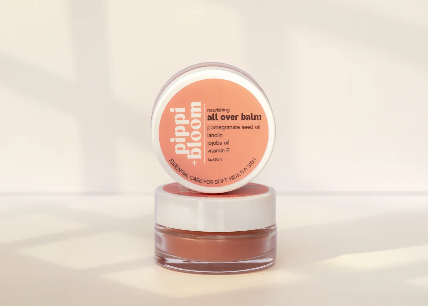 Pippi and Bloom all over balm — one jar open, one lid facing out — in window light against a pale wall