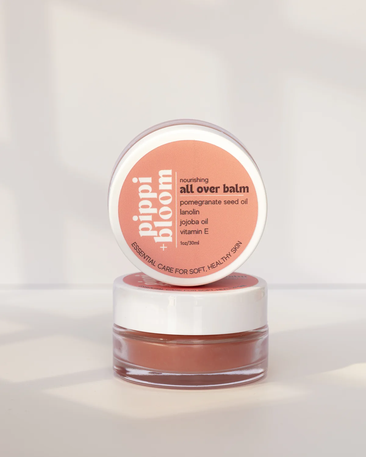 Pippi and Bloom all over balm — one jar open, one lid facing out — in window light against a pale wall