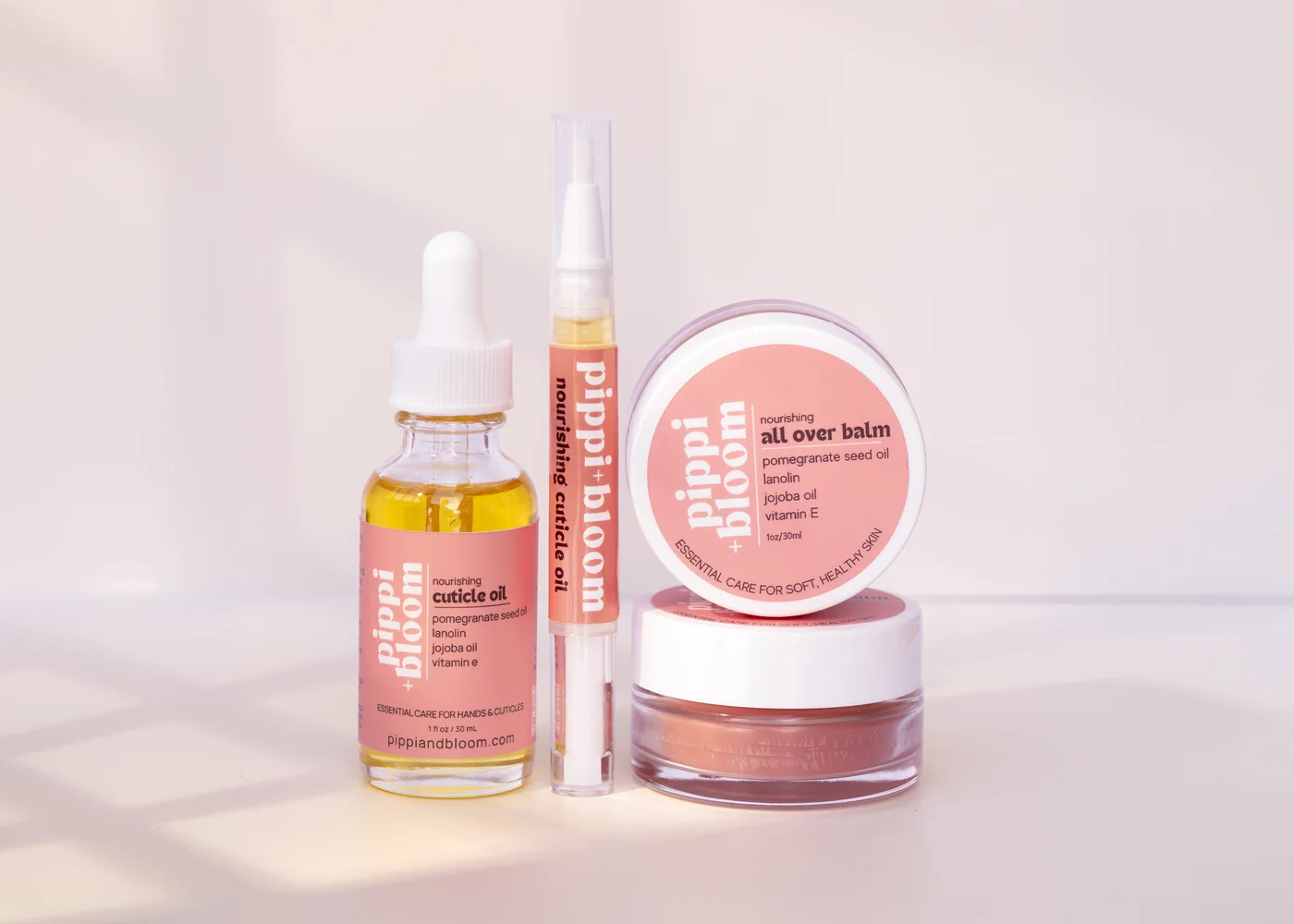 The full Pippi and Bloom line — cuticle oil dropper, brush pen, and all over balm — together in window light against a pale wall